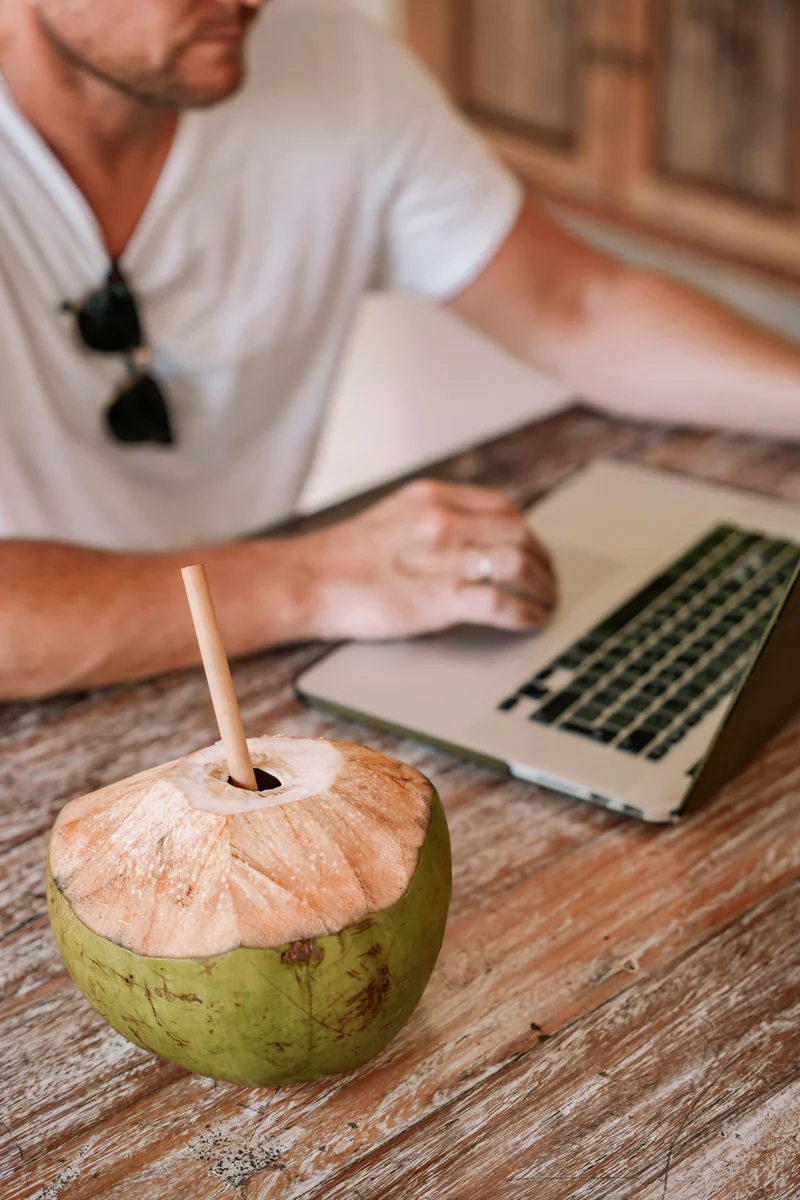 A person working on a laptop beside a fresh coconut at a wooden table